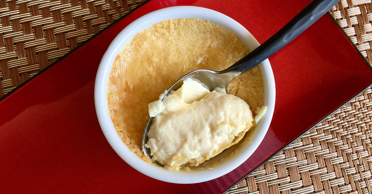 A spoon scoops creamy dessert from a small white dish, revealing a golden-brown top, placed on a red plate with a woven background.