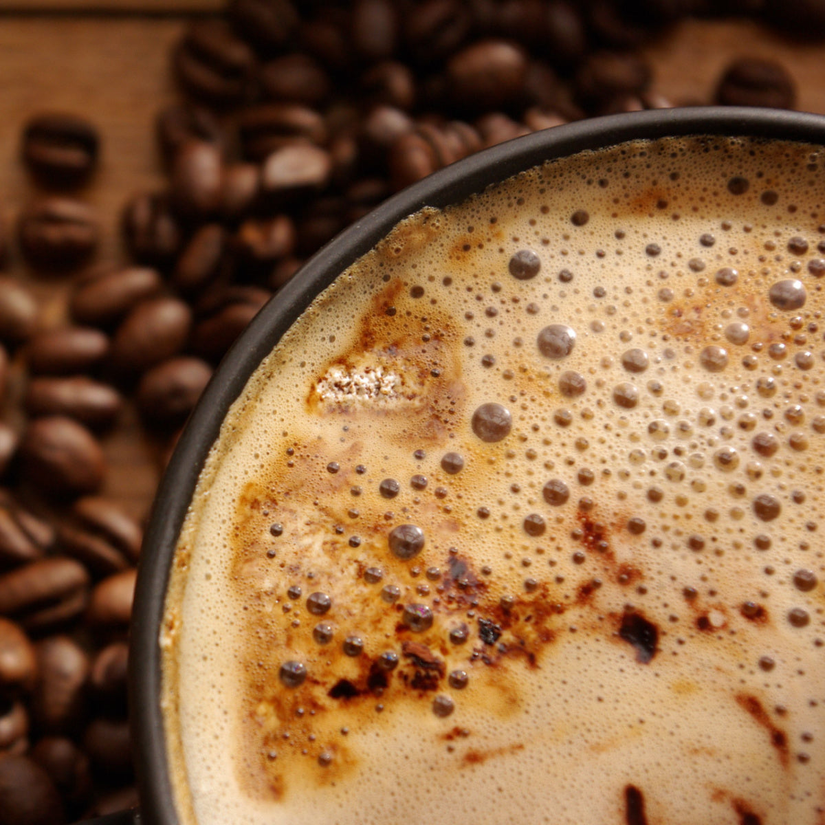 A close-up view of a dark coffee cup filled with frothy coffee, surrounded by scattered coffee beans on a wooden surface.