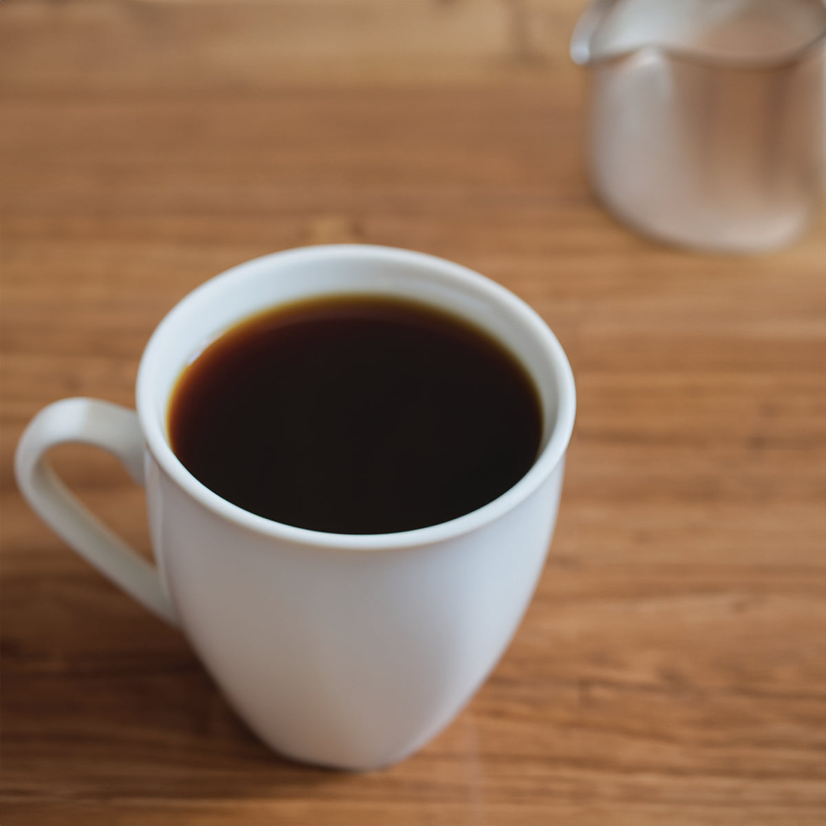 A white mug filled with black coffee on a wooden surface, with a blurred metal creamer in the background.