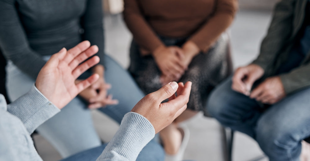 A person gestures with their hand while speaking in a small group discussion, with others sitting nearby, focusing on the conversation in a cozy, informal setting.