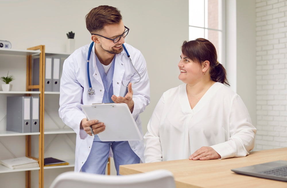 A doctor in a white coat, holding a clipboard, smiles and gestures while discussing with a woman in a white blouse, both seated in a bright, modern office setting.