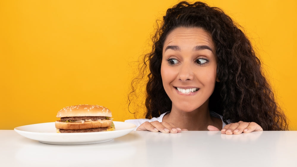 Woman peeking over a table and smiling excitedly at a burger on a plate against a bright yellow background.