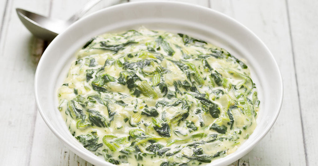 A bowl of creamy spinach with a smooth, rich sauce, served in a white dish on a light wooden surface.