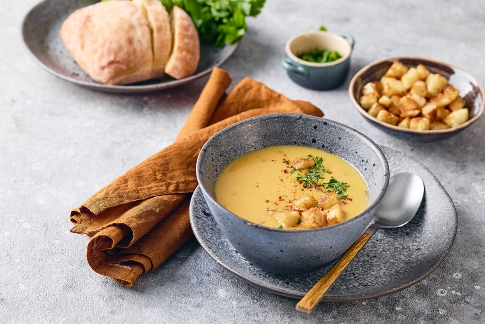 A bowl of creamy soup topped with croutons and herbs is placed on a textured table, accompanied by a brown napkin, a roll of bread, and a small dish of chopped herbs.