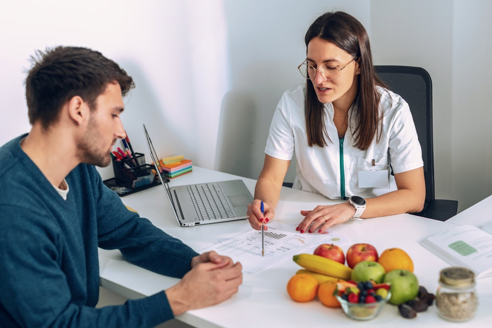 A healthcare professional in a white coat discusses dietary options with a male patient at a desk, surrounded by fresh fruits and a laptop, while pointing to a nutrition plan on paper.