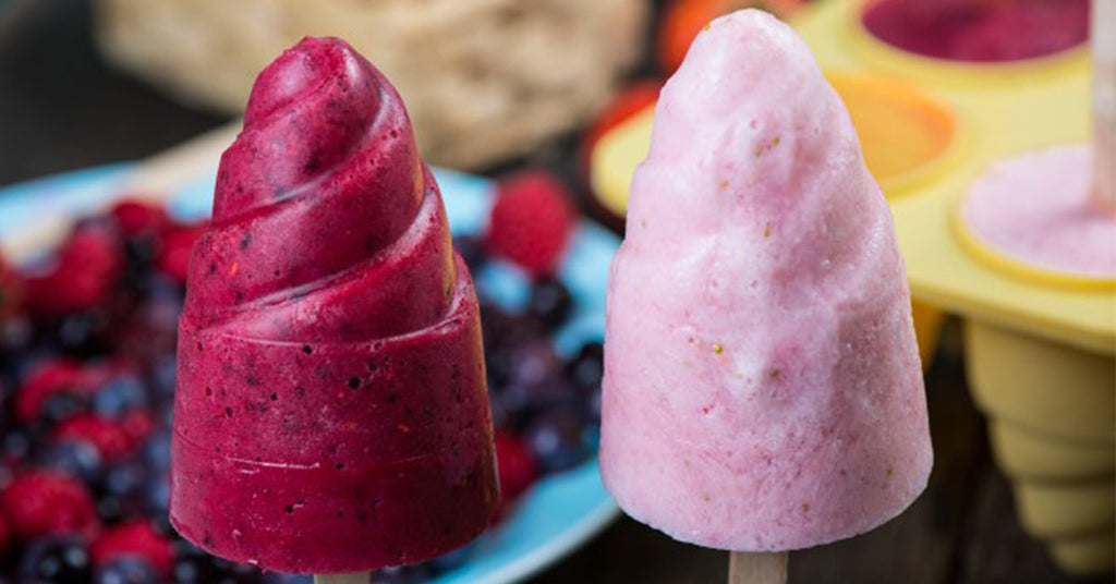 Two colorful frozen pops, one deep red and swirled, the other pale pink and smooth, sit on a wooden table surrounded by fresh berries and a yellow ice pop mold in the background.