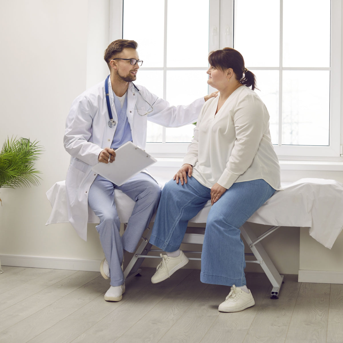 A doctor in a white coat sits on a stool, speaking with a patient in casual clothing who is seated on an exam table, both engaged in a supportive conversation in a bright, modern medical office.