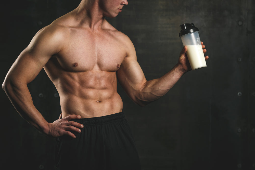 A muscular man in black shorts stands confidently with one hand on his hip, holding a protein shake bottle in the other hand against a dark, gym-like background.
