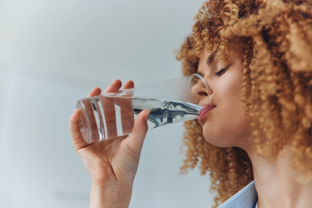 A woman with curly hair drinks water from a clear glass, tilting her head slightly back with her eyes closed, in a bright indoor setting.