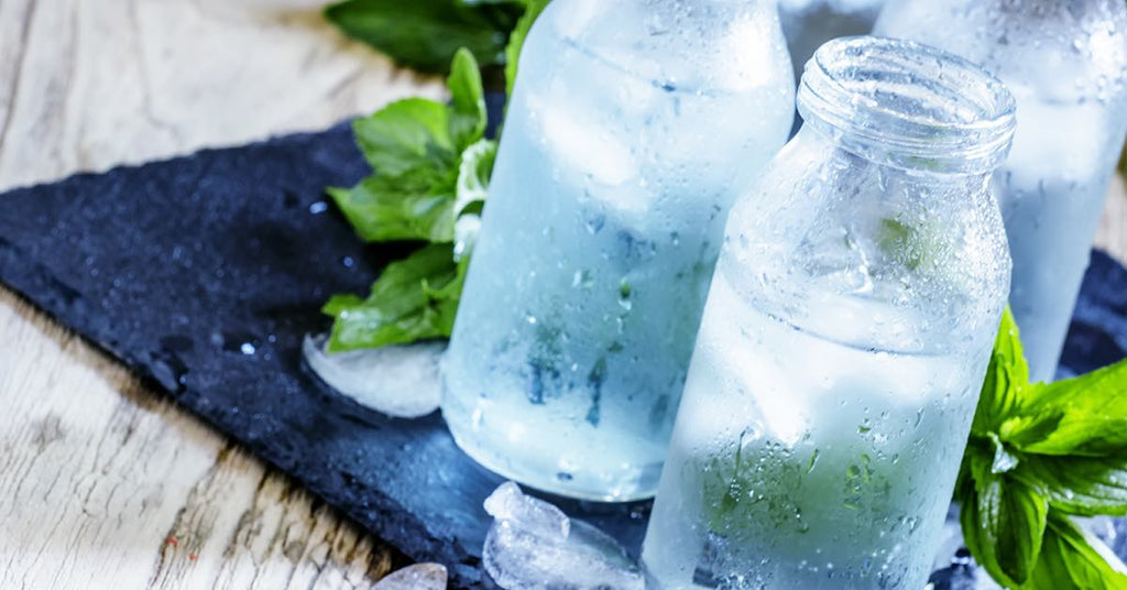Chilled glass bottles filled with ice water and mint leaves, sitting on a slate board with scattered ice.