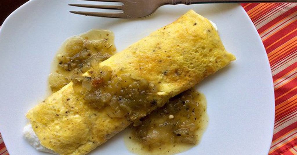 A folded omelet on a white plate topped with a chunky greenish sauce, with a fork placed above it.
