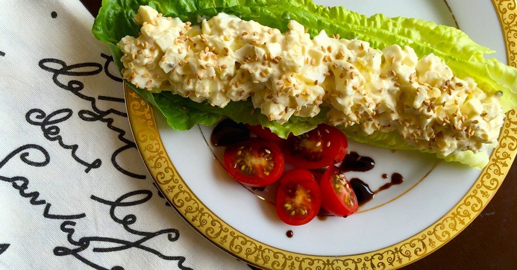 Egg salad on a romaine lettuce leaf with sesame seeds, served with sliced cherry tomatoes and balsamic drizzle.
