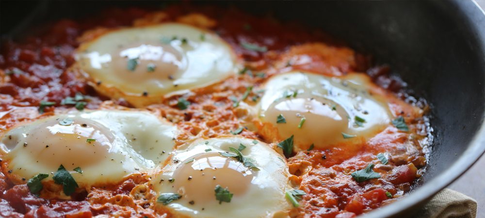 Four sunny-side-up eggs are nestled in a rich, red tomato sauce, garnished with green herbs, all cooked in a black skillet on a wooden surface.
