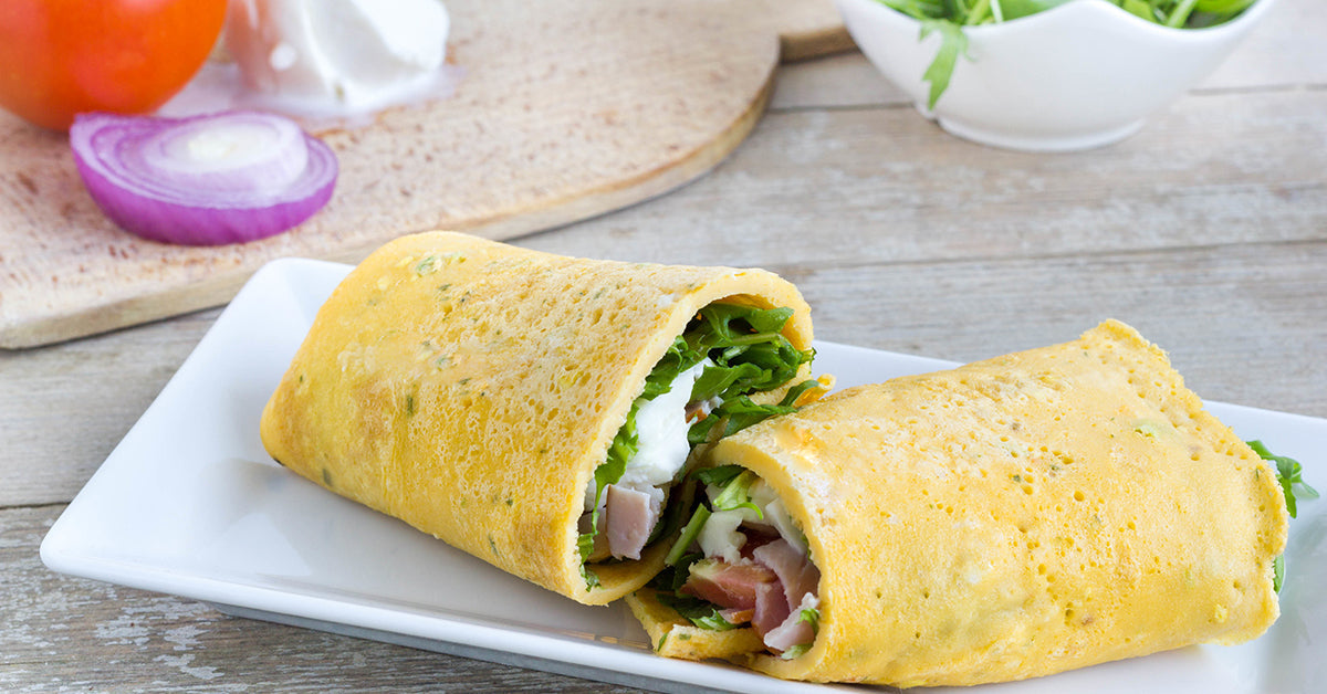 Two egg wraps filled with greens, ham, and cheese on a white plate, with sliced onion and tomato in the background.