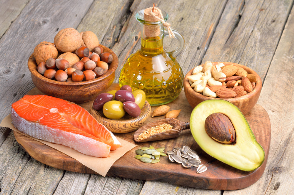 Assortment of healthy fats including salmon, nuts, seeds, olives, avocado, and olive oil on a wooden board.