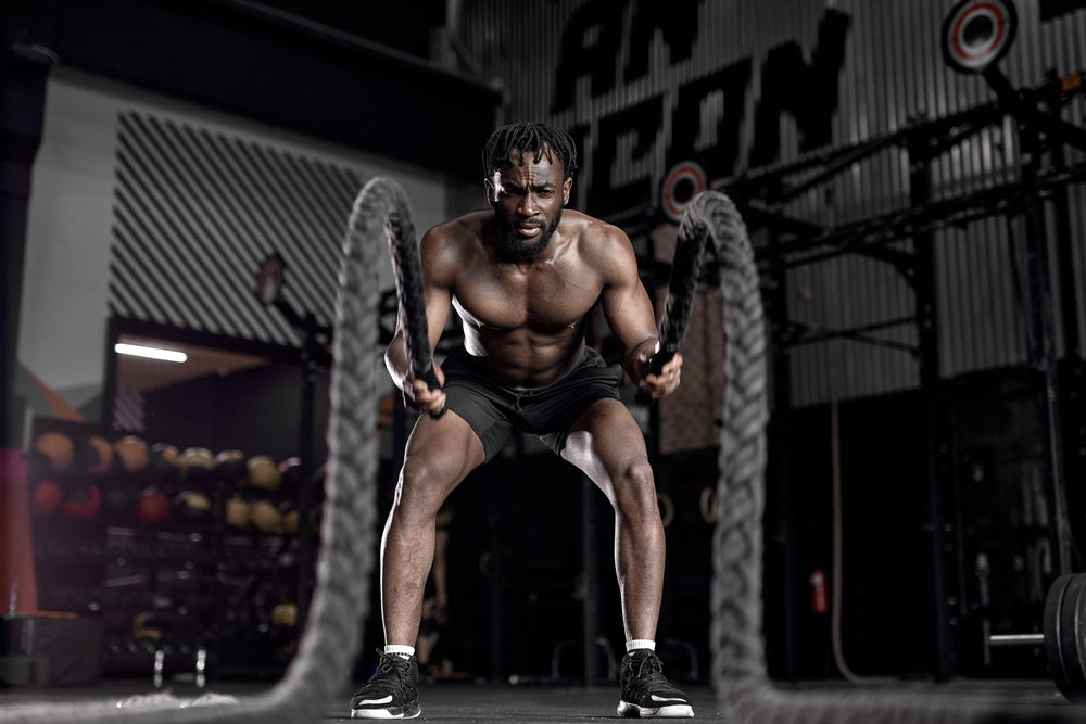 A muscular man is vigorously exercising with battle ropes in a dimly lit gym, focusing intently as he creates waves in the ropes.
