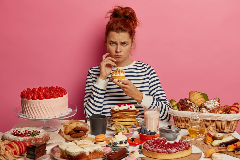A woman with a worried expression sits at a table covered in various desserts and pastries, holding a small cake, against a pink background, reflecting her struggle with food choices.