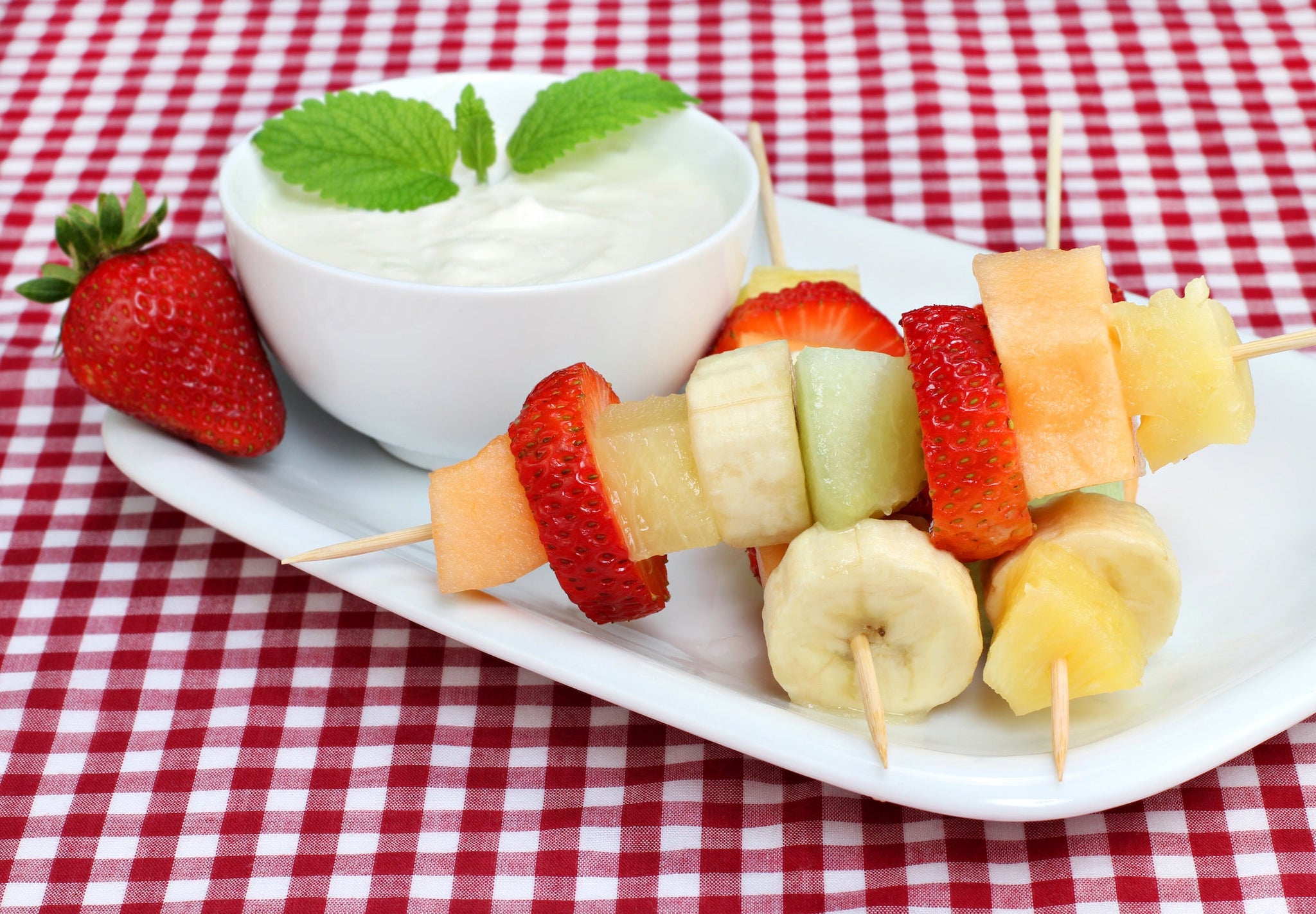Fruit skewers with strawberries, melon, banana, and pineapple served beside a bowl of creamy dip.