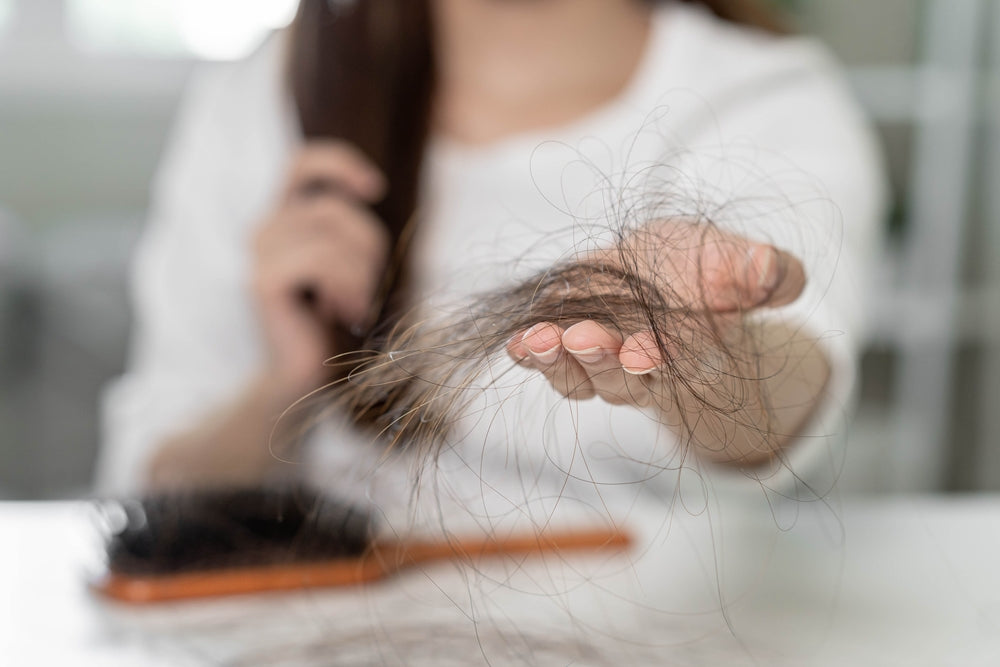 A person in a white shirt holds a clump of hair in their outstretched hand, looking concerned, with a hairbrush resting on a table in a bright indoor setting.