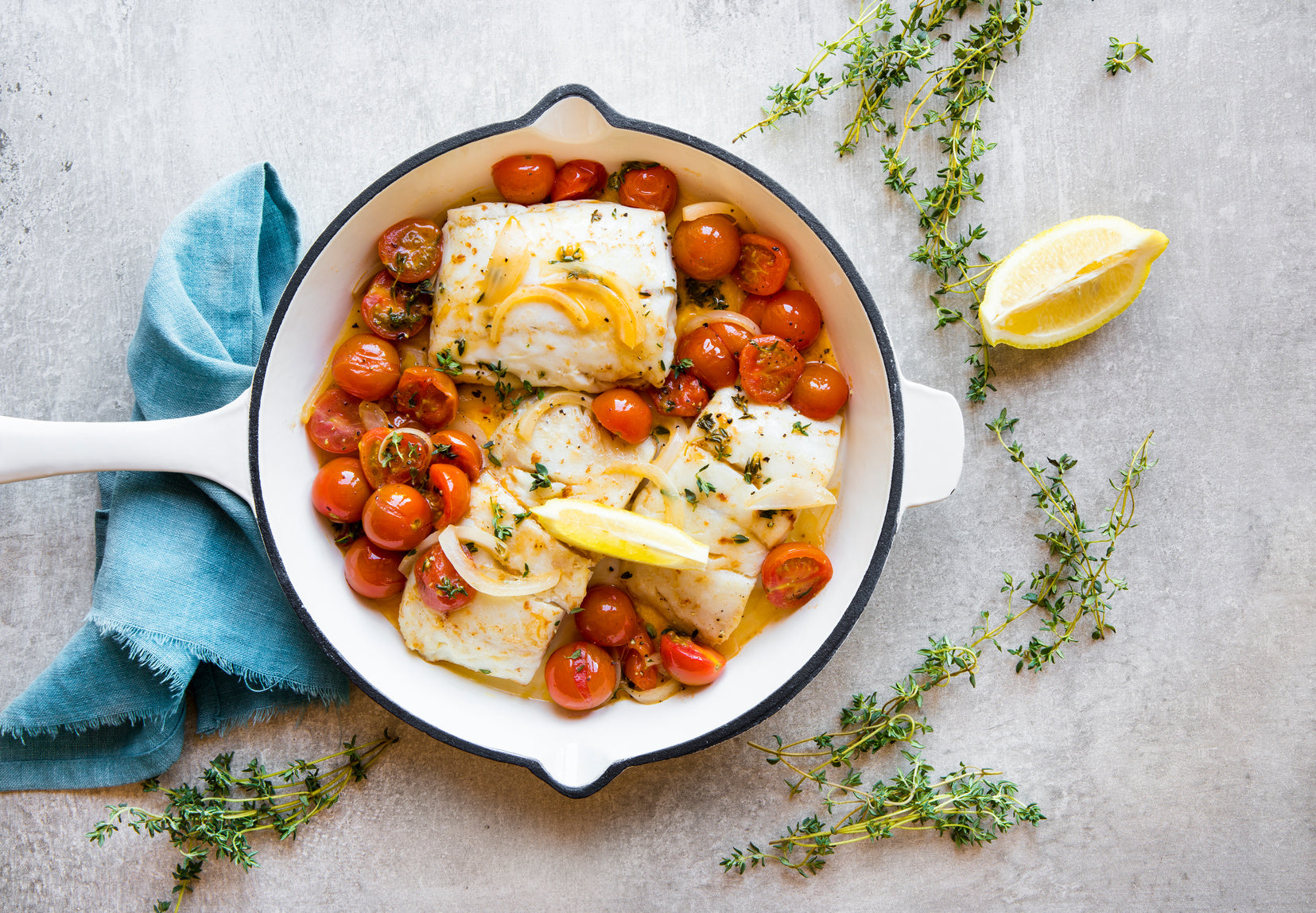 Baked white fish with cherry tomatoes, onions, herbs, and lemon in a white skillet.