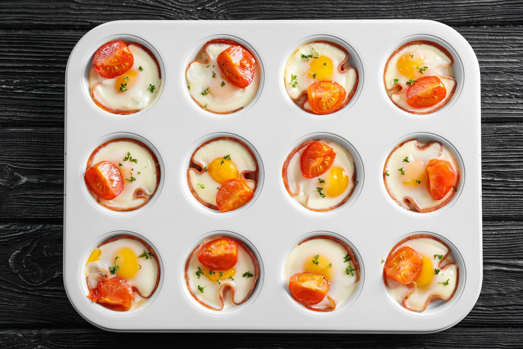 Baked egg cups in a muffin tray with tomatoes and herbs on a dark wooden surface.