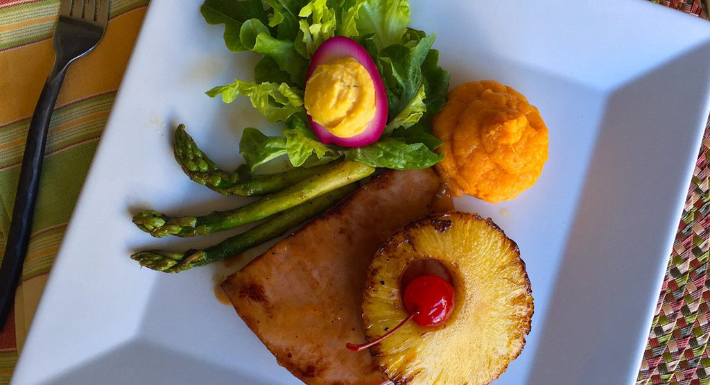Plate with ham topped with pineapple and cherry, asparagus, greens with deviled egg, and mashed sweet potatoes.