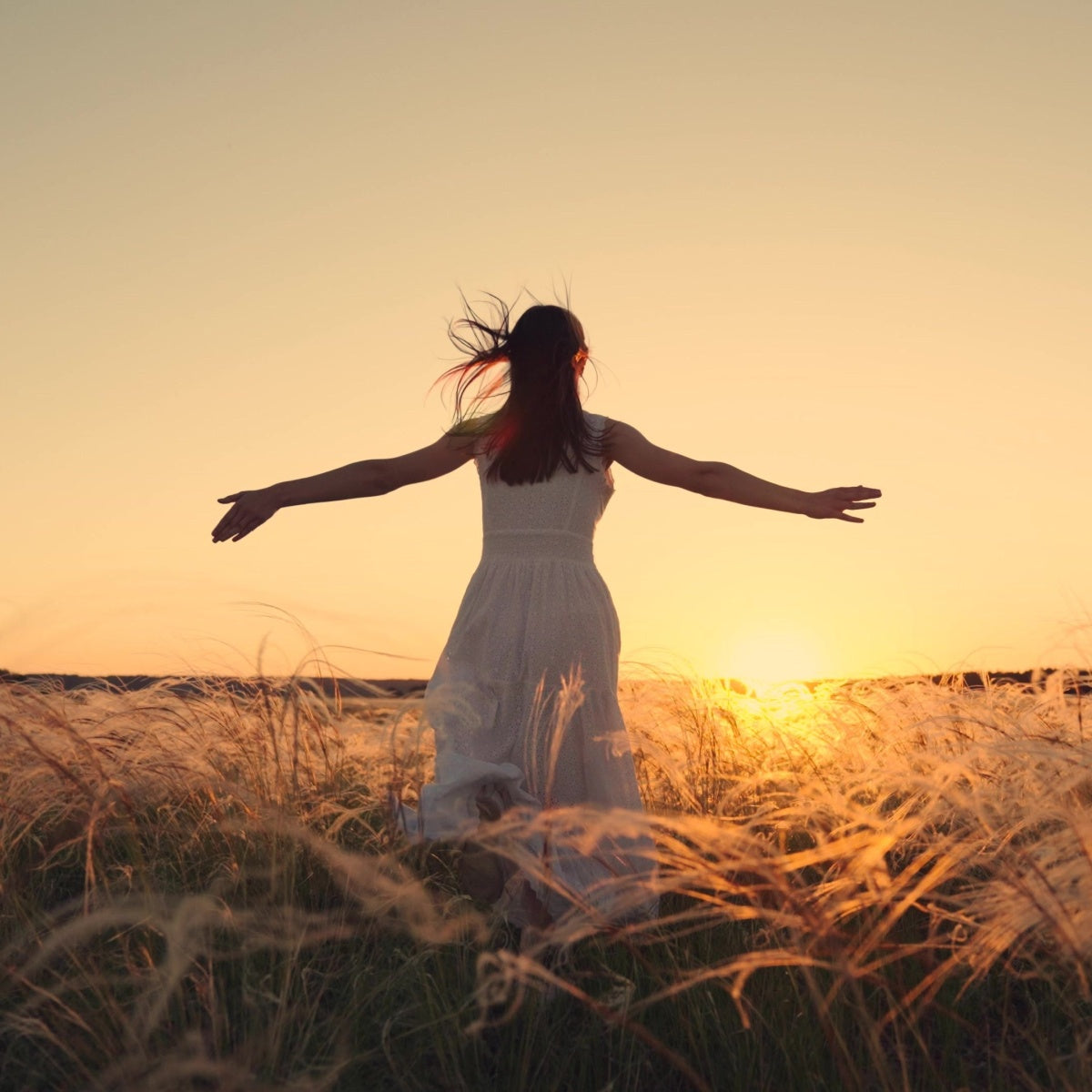 A woman in a flowing white dress stands in a field of tall grass, joyfully spreading her arms as she faces a vibrant sunset, symbolizing freedom and new beginnings after weight loss surgery.