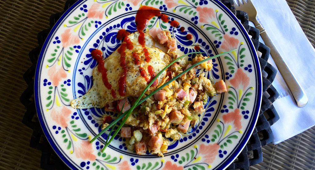 A colorful plate features a fried egg drizzled with ketchup alongside a serving of scrambled eggs mixed with diced ham and vegetables, set on a decorative woven placemat with utensils nearby.
