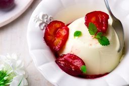 A creamy panna cotta topped with sliced red plums and mint leaves in a white dish.