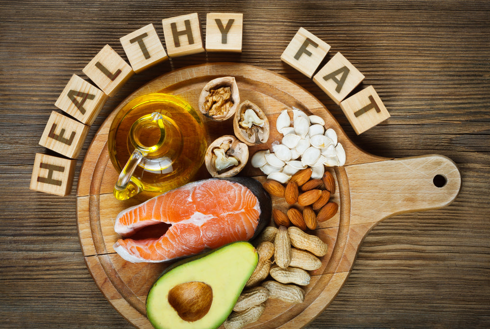 A wooden platter displays a variety of healthy fats, including salmon, avocado, nuts, and olive oil, with wooden blocks spelling "HEALTHY FAT" arranged nearby.