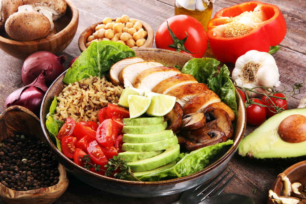 A colorful bowl of grilled chicken, sliced avocado, cherry tomatoes, brown rice, and lettuce is surrounded by fresh vegetables and herbs on a wooden table.