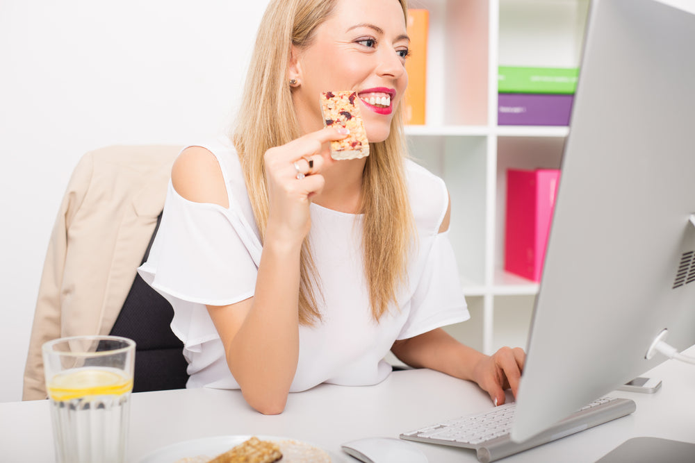 A smiling woman in a white top holds a snack while sitting at a desk with a computer, surrounded by colorful office supplies and a glass of lemon water nearby.