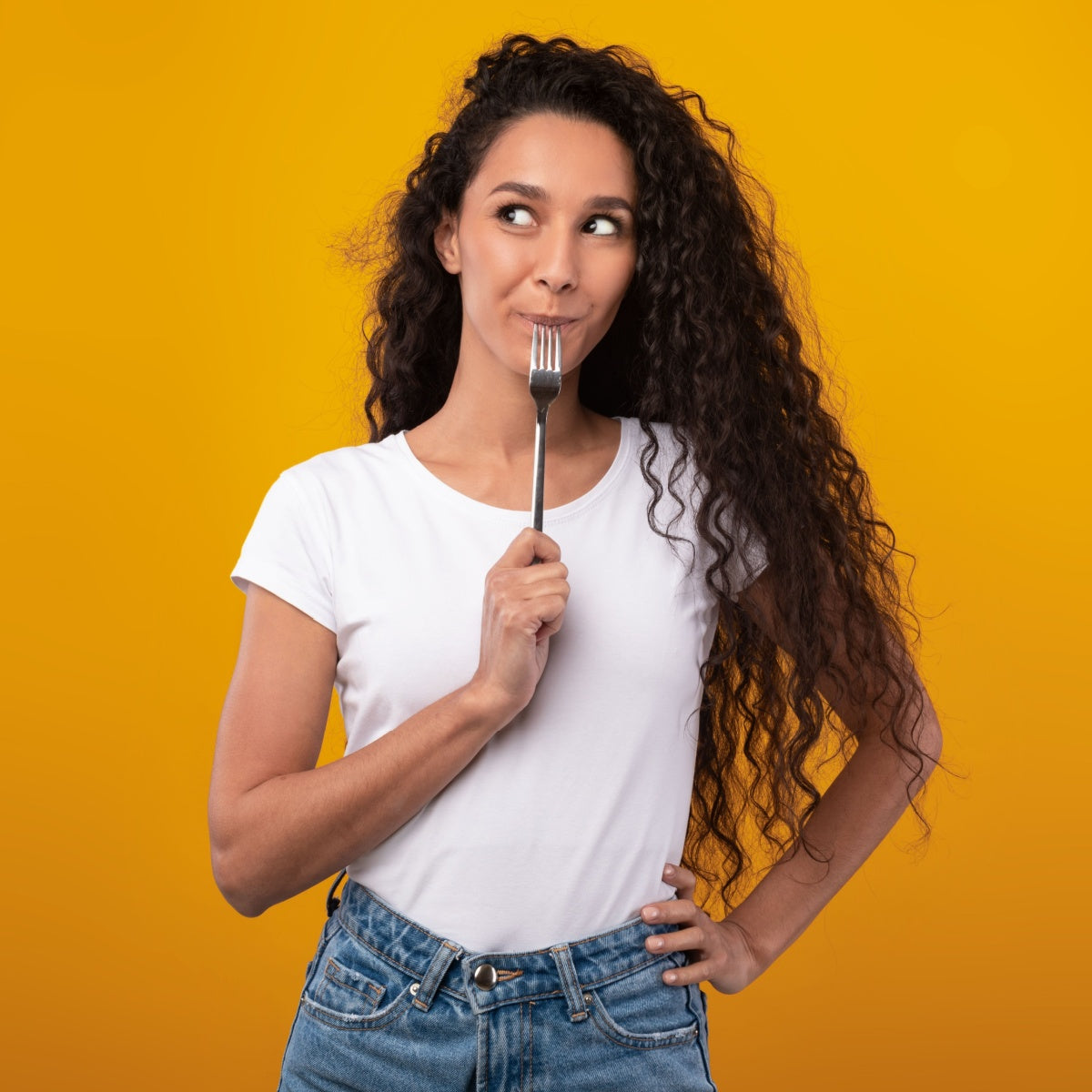 A woman with long, curly hair playfully holds a fork to her lips while looking thoughtfully to the side, set against a bright yellow background, suggesting a lighthearted approach to healthy eating.