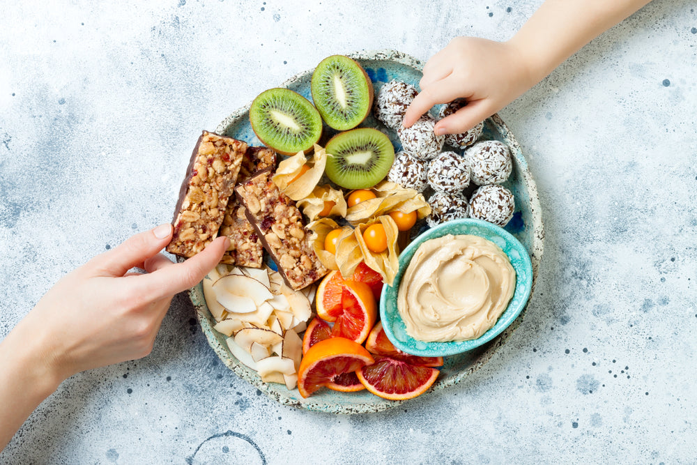 A hand reaches for a granola bar on a colorful platter filled with fresh fruits, coconut slices, energy balls, and a bowl of hummus, set against a light gray background.