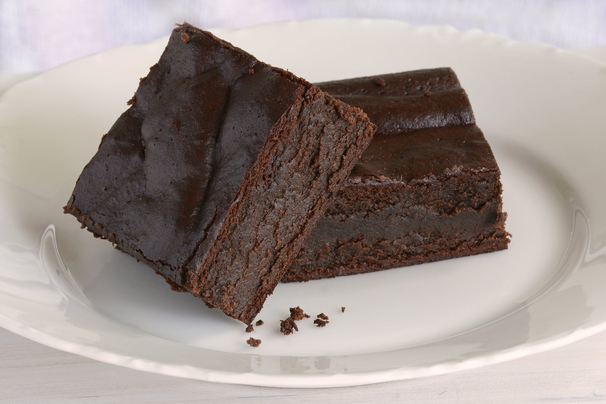 Two rich, dark chocolate brownie squares stacked on a white plate, showing a dense, fudgy texture with a few crumbs.