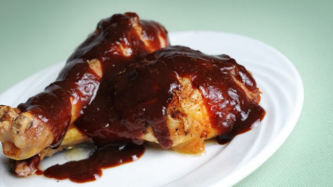 Two pieces of chicken, one drumstick and one thigh, are covered in a glossy barbecue sauce, served on a white plate against a light green background.