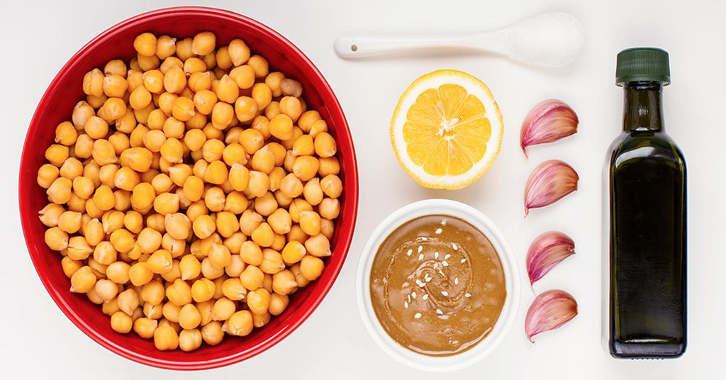 Ingredients for hummus: chickpeas in a red bowl, lemon, garlic cloves, tahini, olive oil, and salt on a white surface.