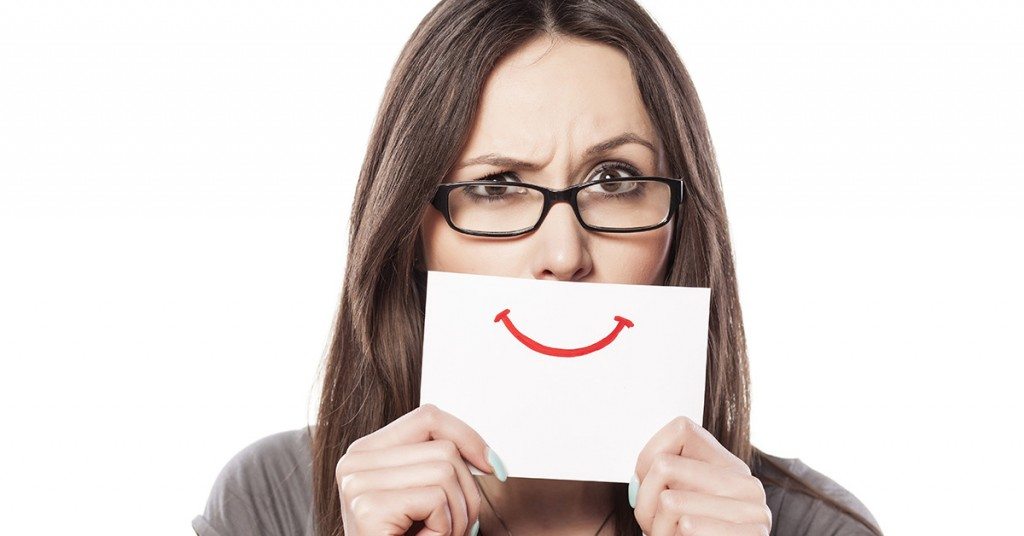 Person wearing glasses holding a card with a drawn smile in front of their mouth, looking concerned or uncertain above it.