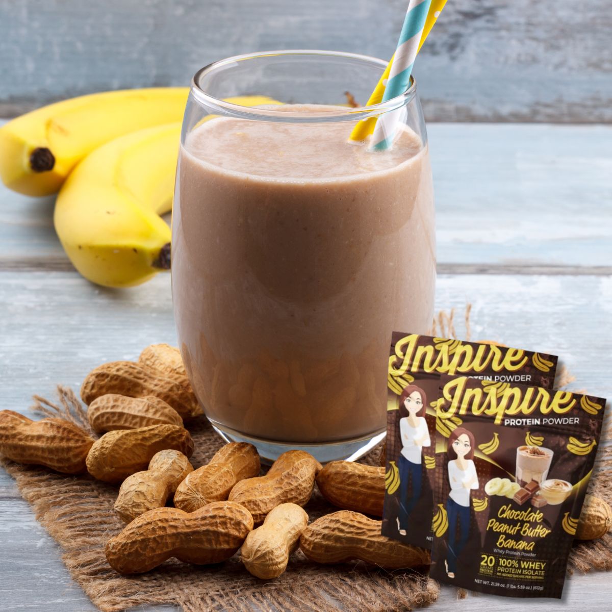 A glass of chocolate peanut butter banana smoothie with two colorful straws is surrounded by peanuts and bananas, alongside a packet of Inspire protein powder on a rustic wooden surface.