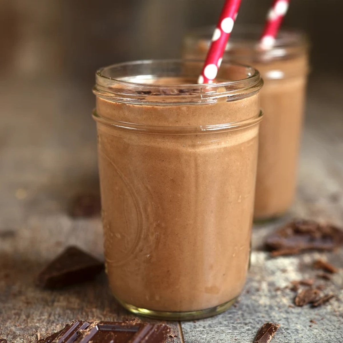 Two jars filled with chocolate smoothie, each with a red and white polka dot straw, sit on a wooden surface alongside pieces of chocolate.