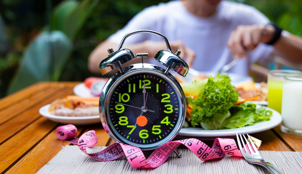 A large alarm clock with bright green numbers sits on a table beside a measuring tape, while a person in a white shirt enjoys a meal of salad and other dishes in a lush outdoor setting.