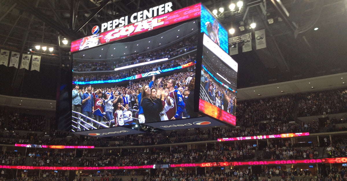 A large crowd cheers and celebrates a goal during a hockey game at the Pepsi Center, with fans wearing team jerseys and an illuminated scoreboard displaying "GOAL."
