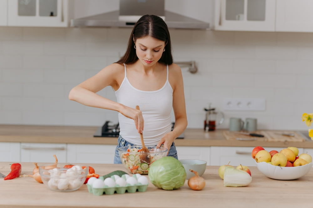A young woman in a white tank top stirs a bowl of salad ingredients at a wooden kitchen counter, surrounded by fresh vegetables, eggs, and fruits, focused on her cooking.