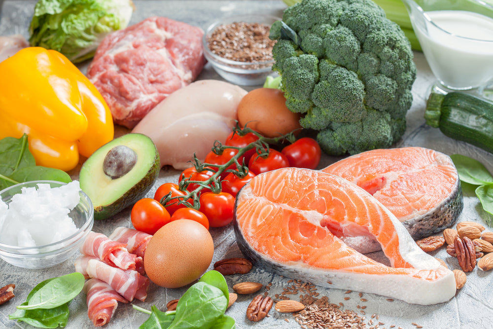 Fresh assorted keto-friendly foods including salmon, meats, vegetables, nuts, eggs, avocado, and tomatoes.