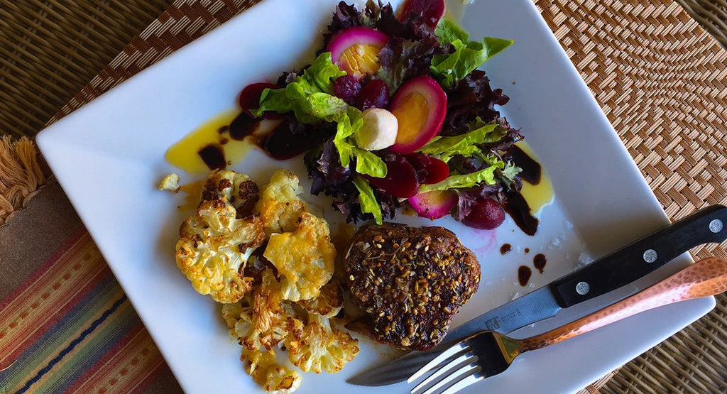 Plate with roasted cauliflower, a seasoned meat patty, and a beet and greens salad drizzled with oil and vinaigrette.