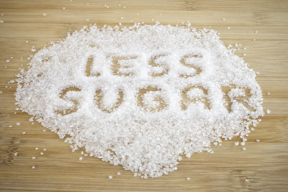 White sugar is scattered on a wooden surface, forming the words "LESS SUGAR" in the center, emphasizing a message about reducing sugar intake.