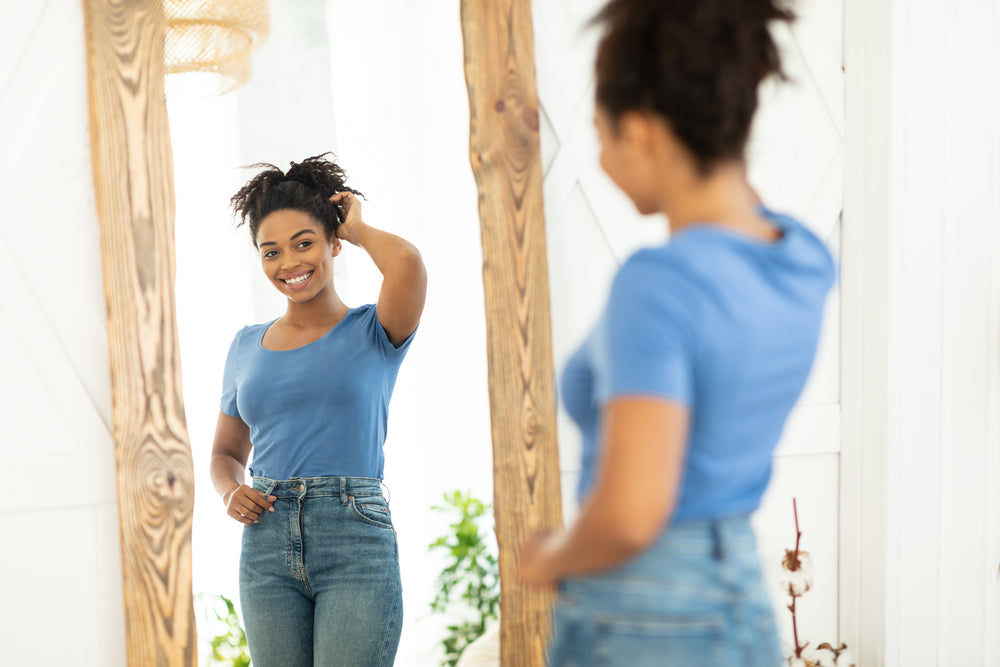 A smiling woman with curly hair stands in front of a mirror, wearing a blue shirt and high-waisted jeans, playfully touching her hair in a bright, airy room with plants in the background.
