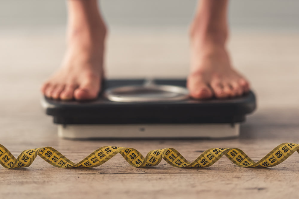 A person stands barefoot on a scale, with a yellow measuring tape coiled on the wooden floor in front, suggesting a focus on weight and body measurements.
