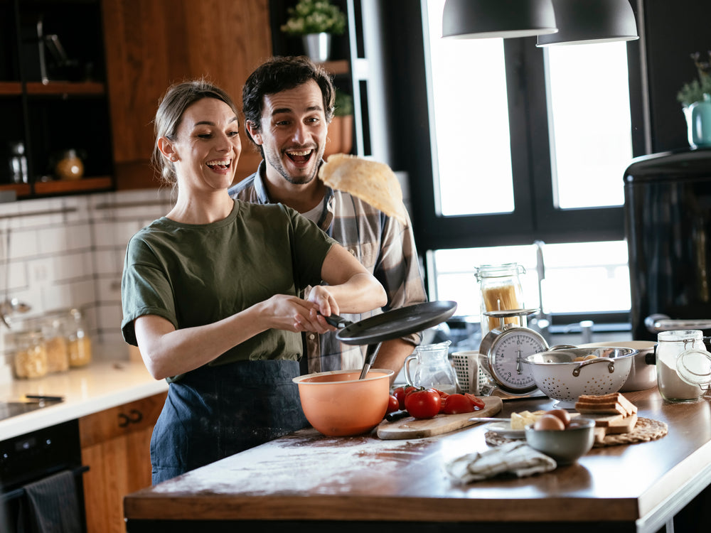 A woman and a man smile joyfully in a modern kitchen as she flips a pancake from a frying pan, with ingredients and cooking tools scattered on the counter around them.