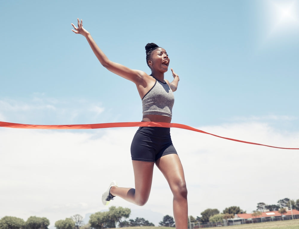 A young woman in athletic gear joyfully crosses the finish line with her arms raised, celebrating her victory on a sunny day at a track and field event.